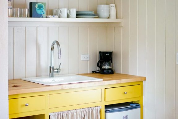 Bright kitchen with a yellow countertop, sink, coffee maker, and dishes on a shelf.